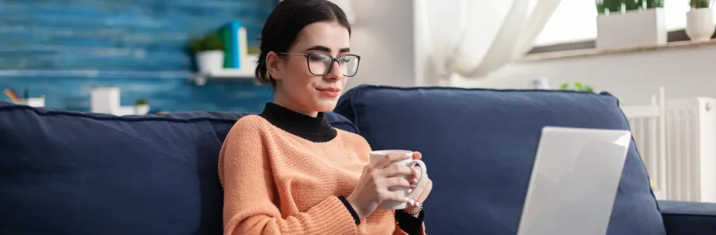 Eine Frau sitzt mit einer Tasse Tee auf der Couch und nimmt an einer Onlineschulung teil.